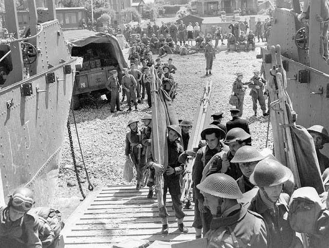 Black and white photograph. Men in battle dress and carrying equipment (such as stretchers) walk towards the camera up the ramp of a landing craft. Behind them, more men in uniform watch as the stand around. A military truck is visible.
